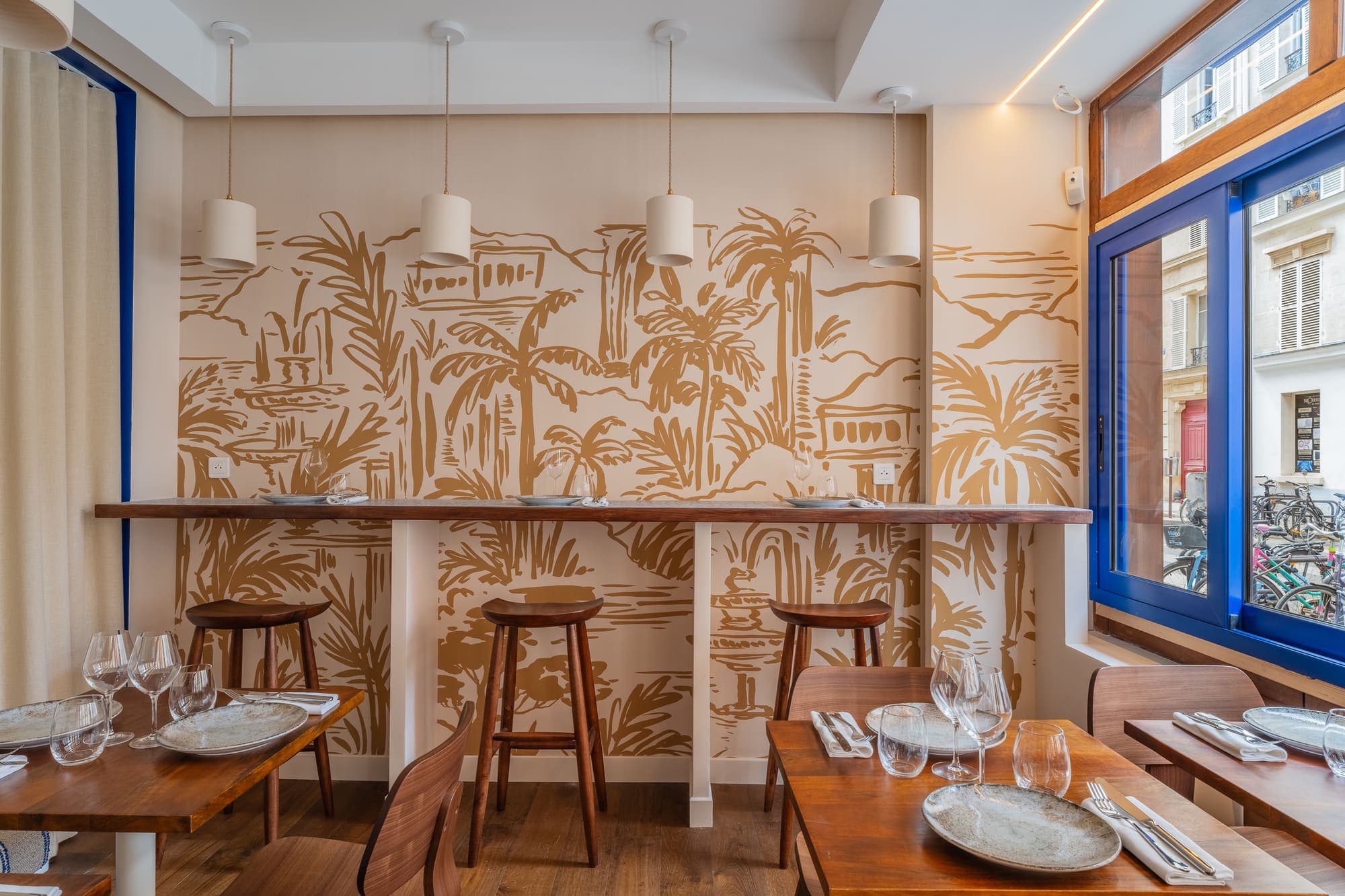 Comptoir haut et tabourets en bois devant une fresque murale de palmiers.