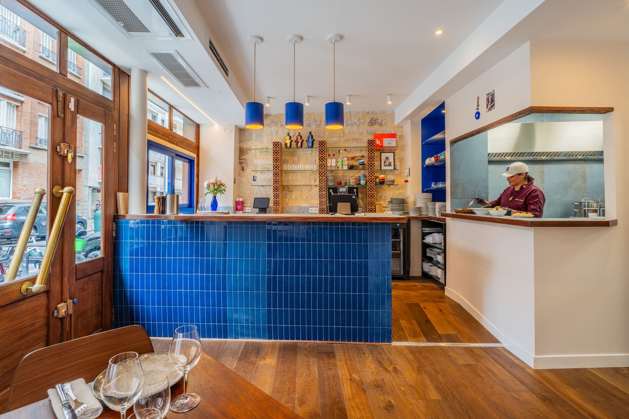 Bar carrelé de bleu avec étagères à bouteilles et cuisine ouverte en fond.