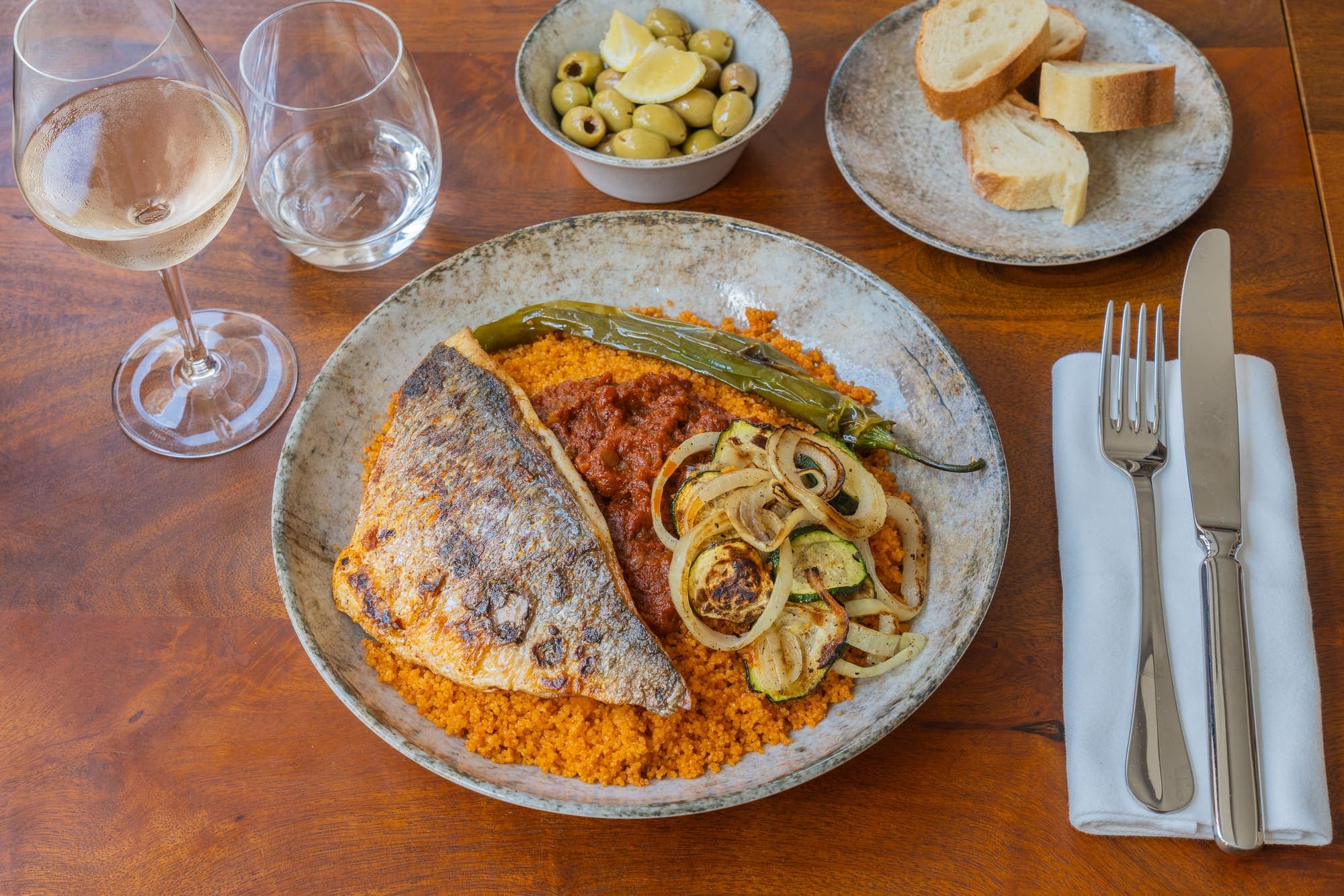 Poisson grillé sur couscous, accompagné d’olives, de pain et d’un verre de vin blanc.