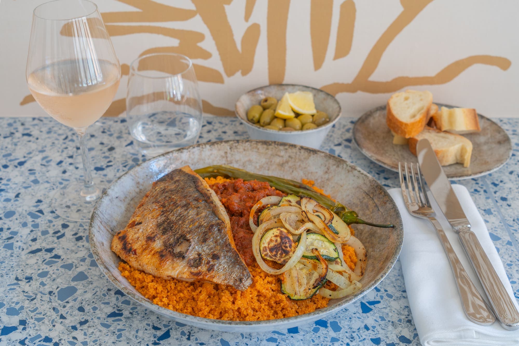 Poisson grillé sur couscous servi devant la fresque murale du restaurant, avec un verre de vin rosé.